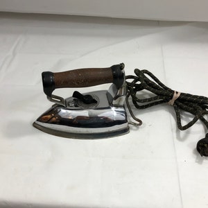 Puede incluir: Una plancha eléctrica vintage de cromo con un mango de madera marrón y un cable negro y verde. La plancha tiene un dial en el lateral para el control de la temperatura.