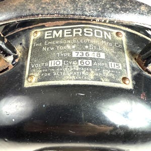 May include: Close-up of a vintage Emerson electric fan. The black metal housing features a rectangular plate with the brand name and manufacturing details. The plate reads "EMERSON" and "NEW YORK * ST. LOUIS".