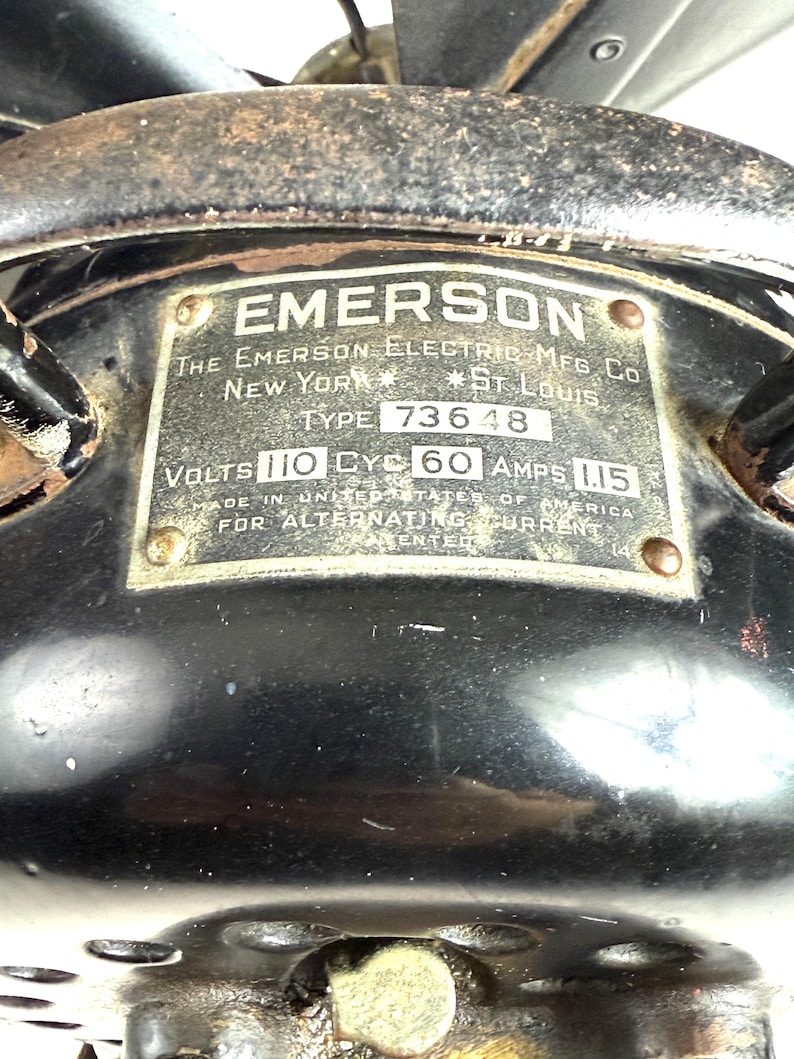 May include: Close-up of a vintage Emerson electric fan. The black metal housing features a rectangular plate with the brand name and manufacturing details. The plate reads "EMERSON" and "NEW YORK * ST. LOUIS".