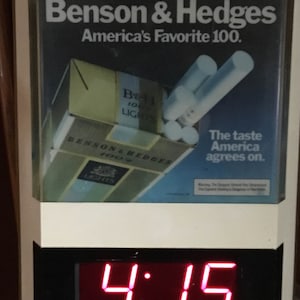 May include: A white digital clock with a red display showing the time 4:15. The clock is framed with a white border and a black border. The clock is advertising Benson & Hedges cigarettes with the text "America's Favorite 100." and "The taste America agrees on."