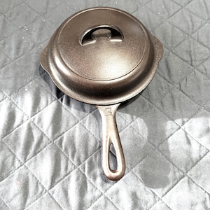 May include: A cast iron skillet with a lid. The skillet is dark brown and has a handle with a hole for hanging. The lid has a small handle on top. The skillet is sitting on a gray quilted surface.