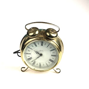 May include: A gold-toned alarm clock with a white face and black Roman numeral hour markers. The clock has a bell-shaped top and a small, black second hand. The clock face reads "Sessions Made in USA".