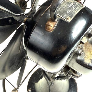 May include: Close-up of a vintage black electric fan. The fan has a shiny black motor housing, curved blades, and a metal cage. A metal plate on the motor reads "The Emerson Electric Mfg. Co. New York".