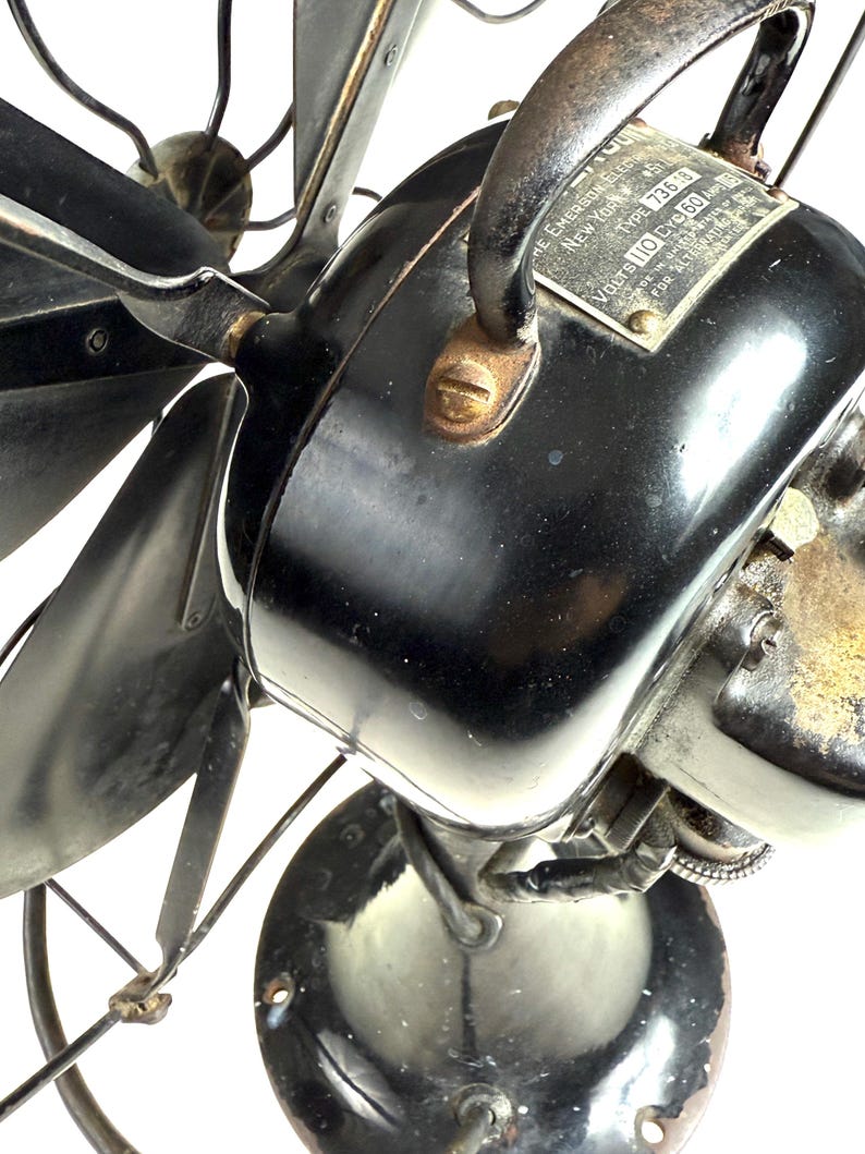 May include: Close-up of a vintage black electric fan. The fan has a shiny black motor housing, curved blades, and a metal cage. A metal plate on the motor reads "The Emerson Electric Mfg. Co. New York".