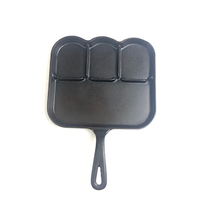 Op de afbeelding: Zwarte gietijzeren koekenpan met een handvat en vier afzonderlijke ronde kookzones.