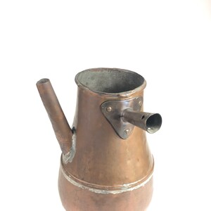 Puede incluir: Cafetera antigua de cobre con un pico y un asa. La cafetera tiene una pátina desgastada y envejecida y una superficie ligeramente abollada.