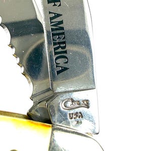 May include: Close-up of a partially open pocket knife. The stainless steel blade is engraved with "B OF AMERICA". The handle is a cream color with a brass pin. The knife has the "Case XX USA" logo and the number "2653" engraved on the blade.