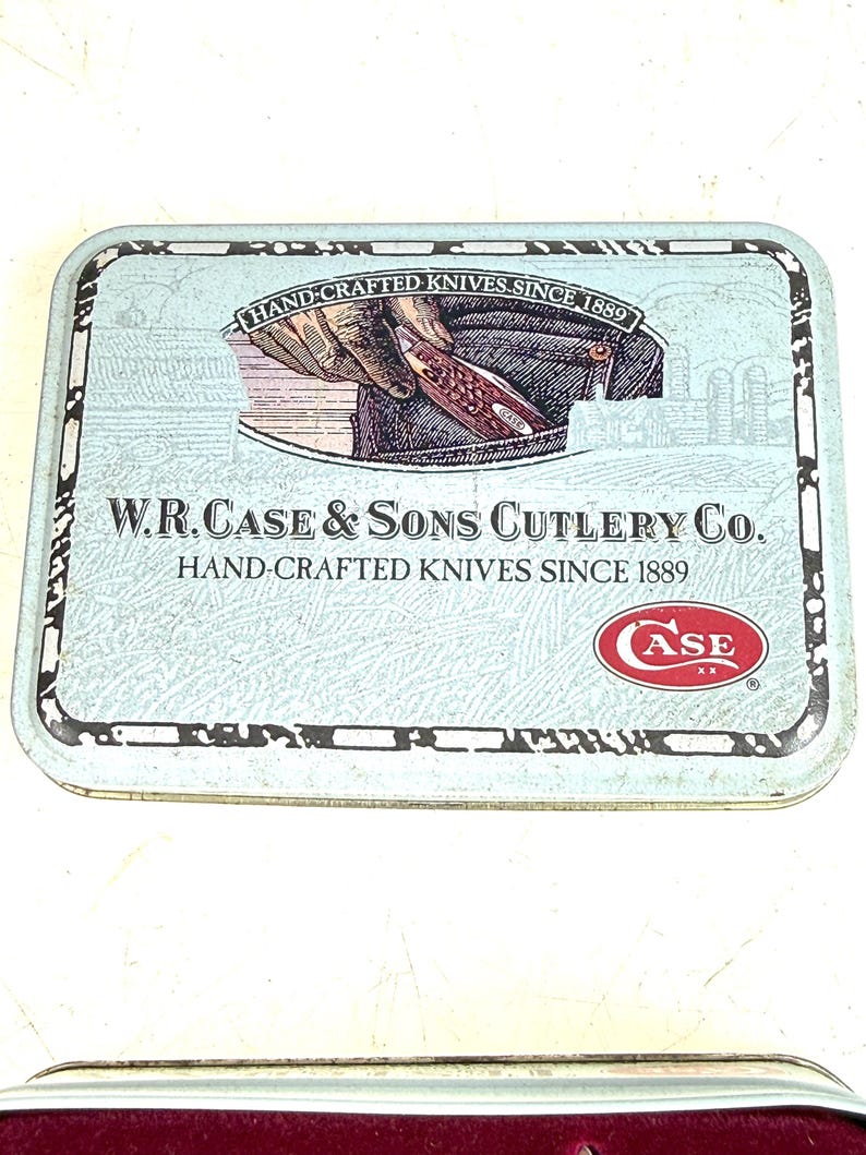 May include: A rectangular, light blue metal tin with the text "HAND-CRAFTED KNIVES SINCE 1889" and "W.R. CASE & SONS CUTLERY CO." The tin features an illustration of a hand holding a knife and the Case logo.