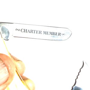May include: A close-up of a silver multi-tool with the text "CHARTER MEMBER" engraved on the blade. The tool has a serrated edge and a handle with a yellow and brown pattern.