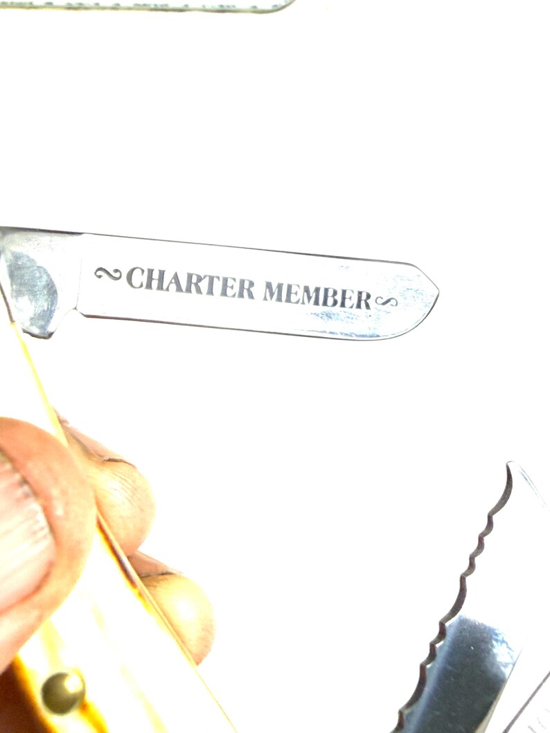 May include: A close-up of a silver multi-tool with the text "CHARTER MEMBER" engraved on the blade. The tool has a serrated edge and a handle with a yellow and brown pattern.
