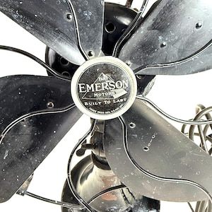 May include: Close-up of a vintage Emerson electric fan. The fan has five black blades and a black metal frame. The center of the fan features a circular emblem with the words "EMERSON MOTORS" and "BUILT TO LAST".