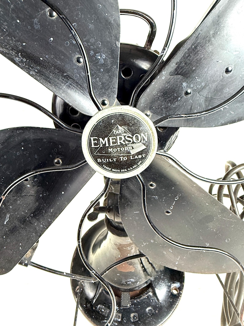 May include: Close-up of a vintage Emerson electric fan. The fan has five black blades and a black metal frame. The center of the fan features a circular emblem with the words "EMERSON MOTORS" and "BUILT TO LAST".