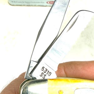 May include: Close-up of a stainless steel pocket knife with a yellow handle. The blade is partially open, revealing the number "6318 SS" etched on the metal. The handle has a brass rivet.