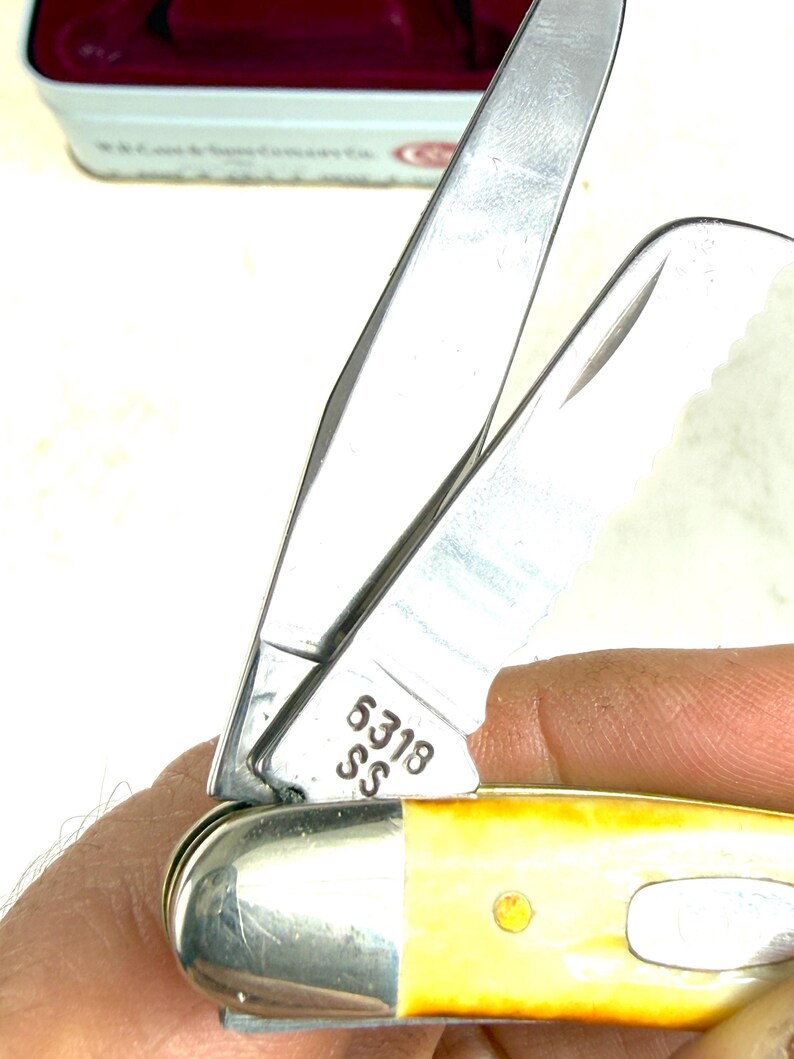 May include: Close-up of a stainless steel pocket knife with a yellow handle. The blade is partially open, revealing the number "6318 SS" etched on the metal. The handle has a brass rivet.