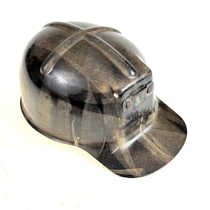 Puede incluir: Casco de seguridad vintage marrón y negro con visera. El casco presenta un patrón texturizado y una pequeña placa metálica en la parte delantera. El casco está diseñado para proteger la cabeza de la caída de objetos.