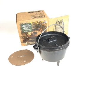May include: A black cast iron camp oven with a lid and a handle. The oven is sitting on three legs and is ready to use. The oven is in a brown cardboard box with the words "Seasoned, Ready to Use Cast Iron Camp Oven" printed on the side. The box also has the words "LODGE LOGIC" printed on the side.