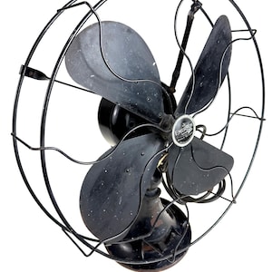 May include: An antique Emerson electric fan with a black metal cage and blades. The fan has a round base and a central motor housing. The blades are slightly curved and the cage has a decorative wire design. The fan is a vintage item.