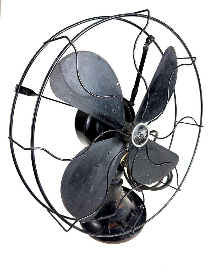 May include: An antique Emerson electric fan with a black metal cage and blades. The fan has a round base and a central motor housing. The blades are slightly curved and the cage has a decorative wire design. The fan is a vintage item.