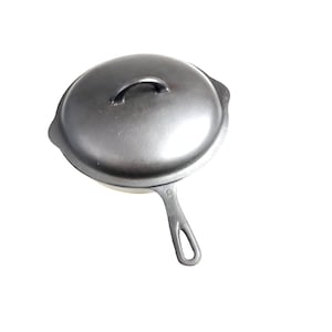 Puede incluir: Una sartén de hierro fundido negra con tapa. La sartén tiene un solo mango y la tapa tiene un pequeño mango en la parte superior.