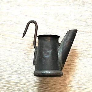 May include: A small, antique, black metal teapot with a curved spout and a handle that forms a hook.