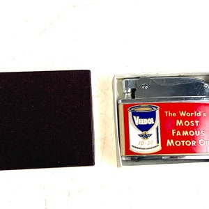 Op de afbeelding: Een zilveren aansteker met een rood en gouden label met het Veedol-logo en de tekst "The World's Most Famous Motor Oil". De aansteker zit in een witte doos.