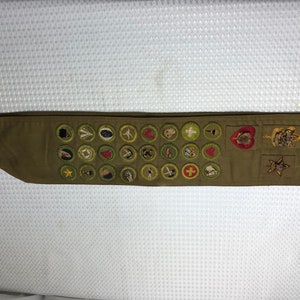May include: A brown Boy Scout sash with a variety of merit badges, including a heart, a cross, and a star. The sash is decorated with a variety of colors, including green, red, and yellow.