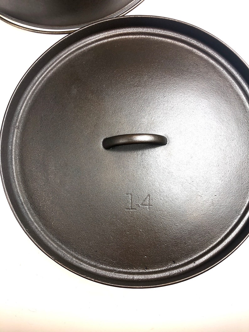 Puede incluir: Tapa de olla de hierro fundido negro con asa y el n&uacute;mero "14" inscrito en la parte superior.