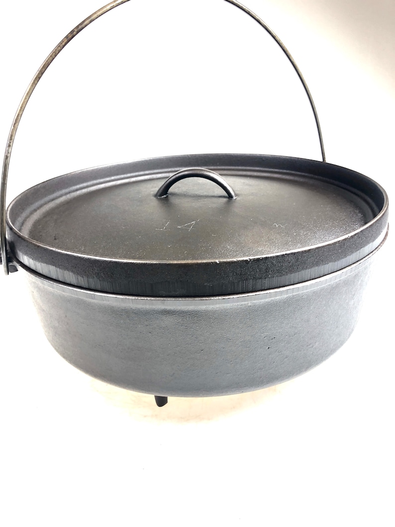 Puede incluir: Una olla de hierro fundido negra con un asa de alambre y tres patas. La tapa tiene una peque&ntilde;a asa y el n&uacute;mero "14" est&aacute; inscrito en la tapa.