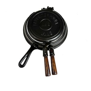 Może przedstawiać: Czarne żeliwne żelazko do gofrów z drewnianym uchwytem. Żelazko do gofrów ma napis "Favorite Pie Iron No. 8 Ware" na górze.