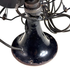 May include: Close-up of a vintage black electric fan. The fan has a rounded base, a slender neck, and a cage protecting the blades. The fan's components are black, with some signs of wear and tear, and the blades are visible.