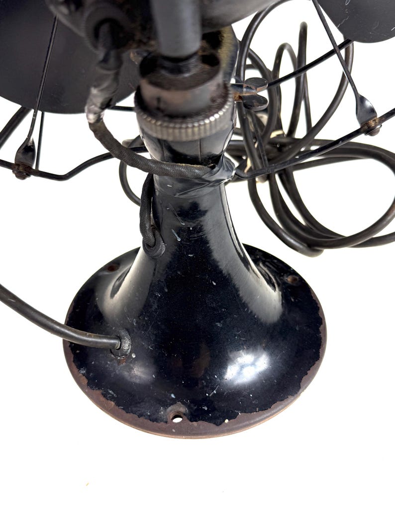 May include: Close-up of a vintage black electric fan. The fan has a rounded base, a slender neck, and a cage protecting the blades. The fan's components are black, with some signs of wear and tear, and the blades are visible.