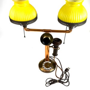 May include: An antique telephone lamp with two yellow glass shades. The lamp features a copper and brass base, a rotary dial, and a handset. The shades have a ribbed design and are trimmed with a decorative metal edge.
