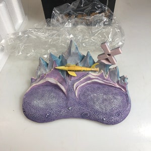 May include: A purple and blue ceramic sculpture of a Martian landscape with a yellow rocket ship and a cross on top. The rocket ship has the words "MARTIAN MARAUDER" printed on it.