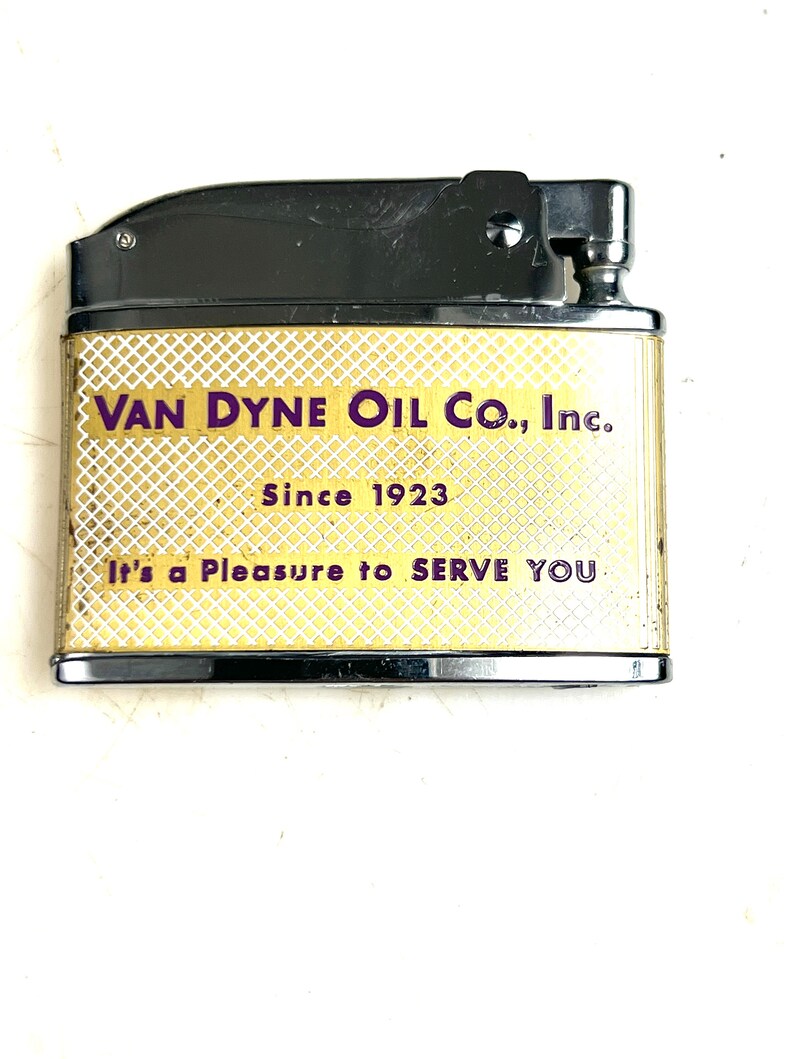 Op de afbeelding: Een goudkleurige aansteker met een zwarte bovenkant en een wit en gouden ruitpatroon. De aansteker heeft de tekst "VAN DYNE OIL CO., Inc. Since 1923 It's a Pleasure to SERVE YOU" erop gedrukt.