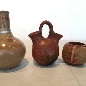 Może przedstawiać: Trzy ręcznie robione naczynia ceramiczne. Okrągły wazon w jasnobrązowym kolorze z szerokim otworem i białym paskiem wokół szyjki. Czerwono-brązowy dzbanek z dwoma wylewkami i zaokrąglonym korpusem. Mała, okrągła, czerwono-brązowa miska z geometrycznymi wzorami.