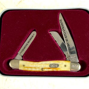 May include: A Case XX pocket knife with a cream-colored handle and two open blades. One blade reads "HANDYMAN CLUB OF AMERICA," and the other reads "CHARTER MEMBER." The knife is displayed in a red velvet-lined tin.
