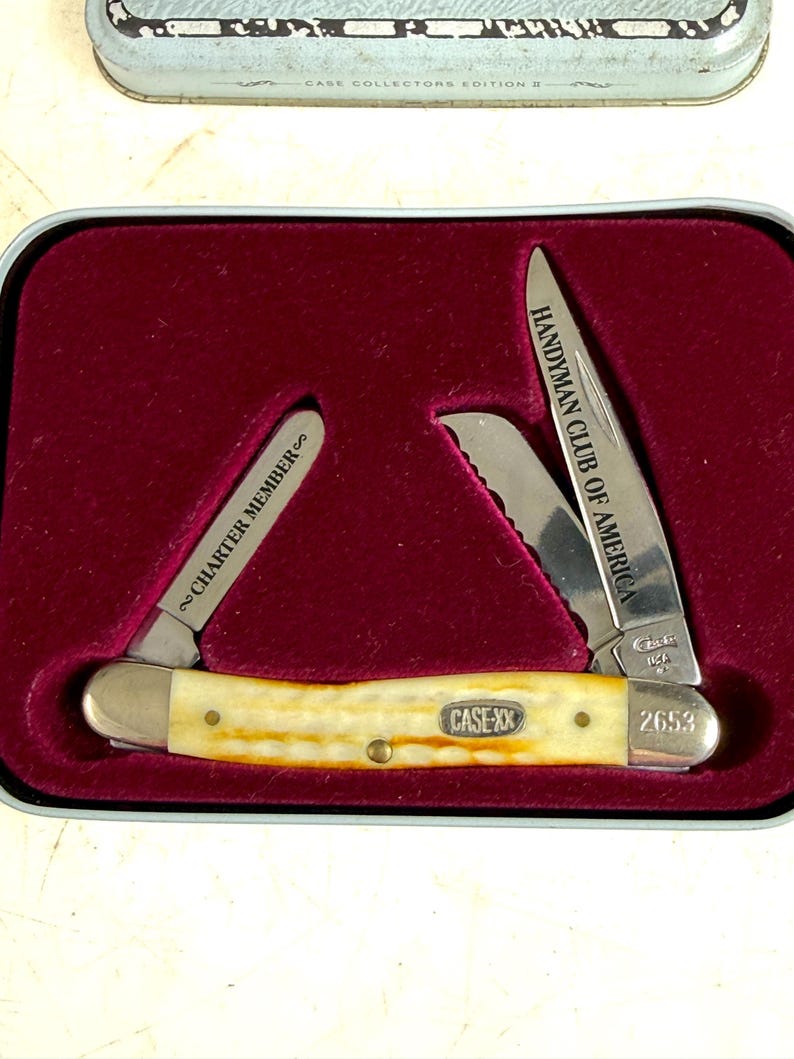 May include: A Case XX pocket knife with a cream-colored handle and two open blades. One blade reads "HANDYMAN CLUB OF AMERICA," and the other reads "CHARTER MEMBER." The knife is displayed in a red velvet-lined tin.