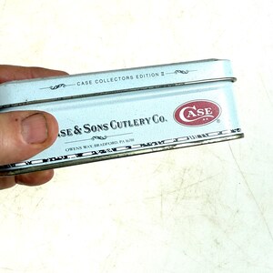 May include: A light blue metal box with the text "CASE COLLECTORS EDITION II" and "CASE & SONS CUTLERY CO." in black. The Case logo is in red. The box is held by a hand.