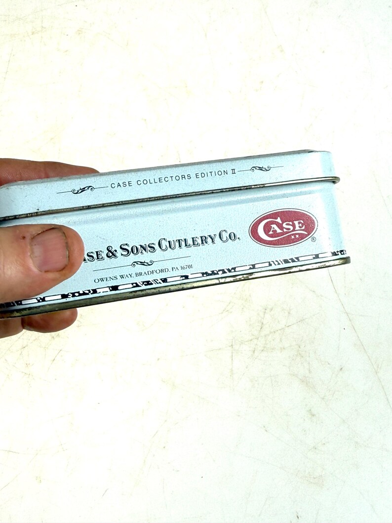 May include: A light blue metal box with the text "CASE COLLECTORS EDITION II" and "CASE & SONS CUTLERY CO." in black. The Case logo is in red. The box is held by a hand.