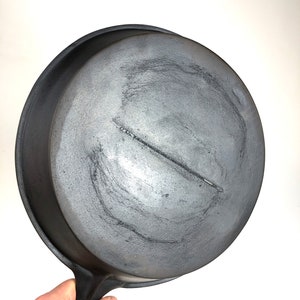 May include: A black cast iron skillet with a smooth, slightly worn surface. The skillet has a single handle and is being held by a hand.