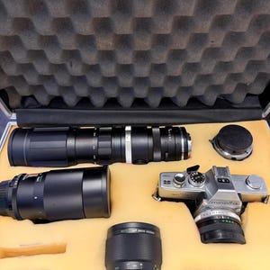 May include: A vintage Minolta SR-7 camera with multiple lenses and accessories, including a long telephoto lens, macro lens, and lens caps. The camera and lenses are stored in a protective case with yellow foam inserts.