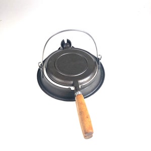 Może przedstawiać: Czarne żeliwne żelazko do gofrów z drewnianym uchwytem i metalowym uchwytem do przenoszenia. Żelazko do gofrów jest zamknięte i gotowe do użycia.