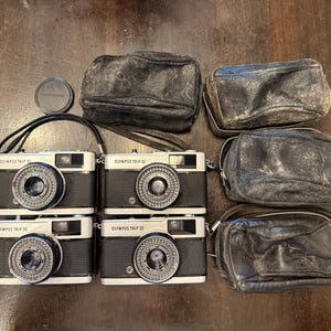 Op de afbeelding: Een verzameling Olympus Trip 35 camera's met zwarte en zilveren behuizingen, samen met verschillende versleten zwarte leren etuis en een lensdop. De camera's hebben opvallende lenzen en de tekst "OLYMPUS TRIP 35" is zichtbaar.