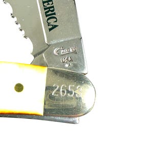 May include: Close-up of a partially open pocket knife. The blade is silver with a serrated edge. The handle is white with a brass rivet. The knife has the text "OF AMERICA" and "2653" on the blade.