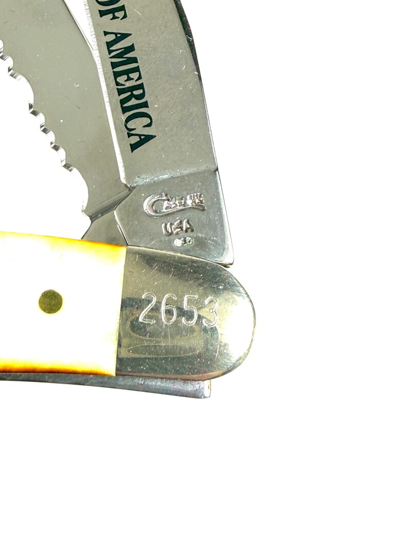 May include: Close-up of a partially open pocket knife. The blade is silver with a serrated edge. The handle is white with a brass rivet. The knife has the text "OF AMERICA" and "2653" on the blade.
