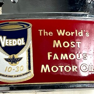 Op de afbeelding: Een vintage aansteker met een rood en gouden ontwerp met een blikje Veedol motorolie. De tekst op de aansteker luidt "The World's Most Famous Motor Oil".