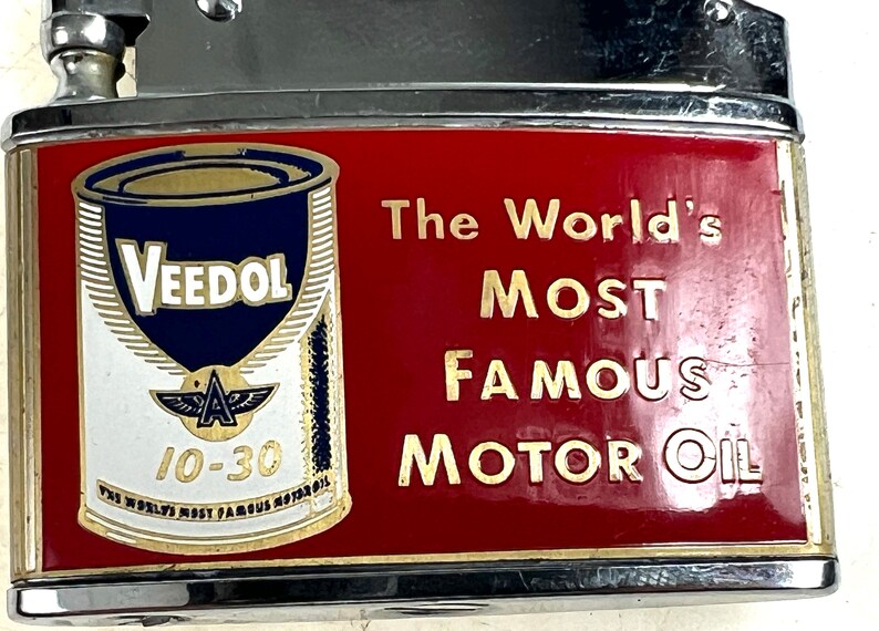 Op de afbeelding: Een vintage aansteker met een rood en gouden ontwerp met een blikje Veedol motorolie. De tekst op de aansteker luidt "The World's Most Famous Motor Oil".