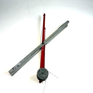 May include: A vintage red fishing rod with a silver reel and a grey wooden crossbar attached. The reel features circular text that reads THE FISHERMANS PAL and includes the text BOX 804. The rod is positioned vertically with the crossbar angled across it.