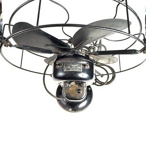 May include: An antique black electric fan with three blades, enclosed in a protective wire cage. The fan's motor housing is also black, with a small metal plate. The fan is designed to be mounted on a wall.
