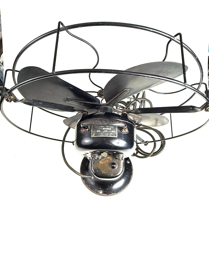 May include: An antique black electric fan with three blades, enclosed in a protective wire cage. The fan's motor housing is also black, with a small metal plate. The fan is designed to be mounted on a wall.